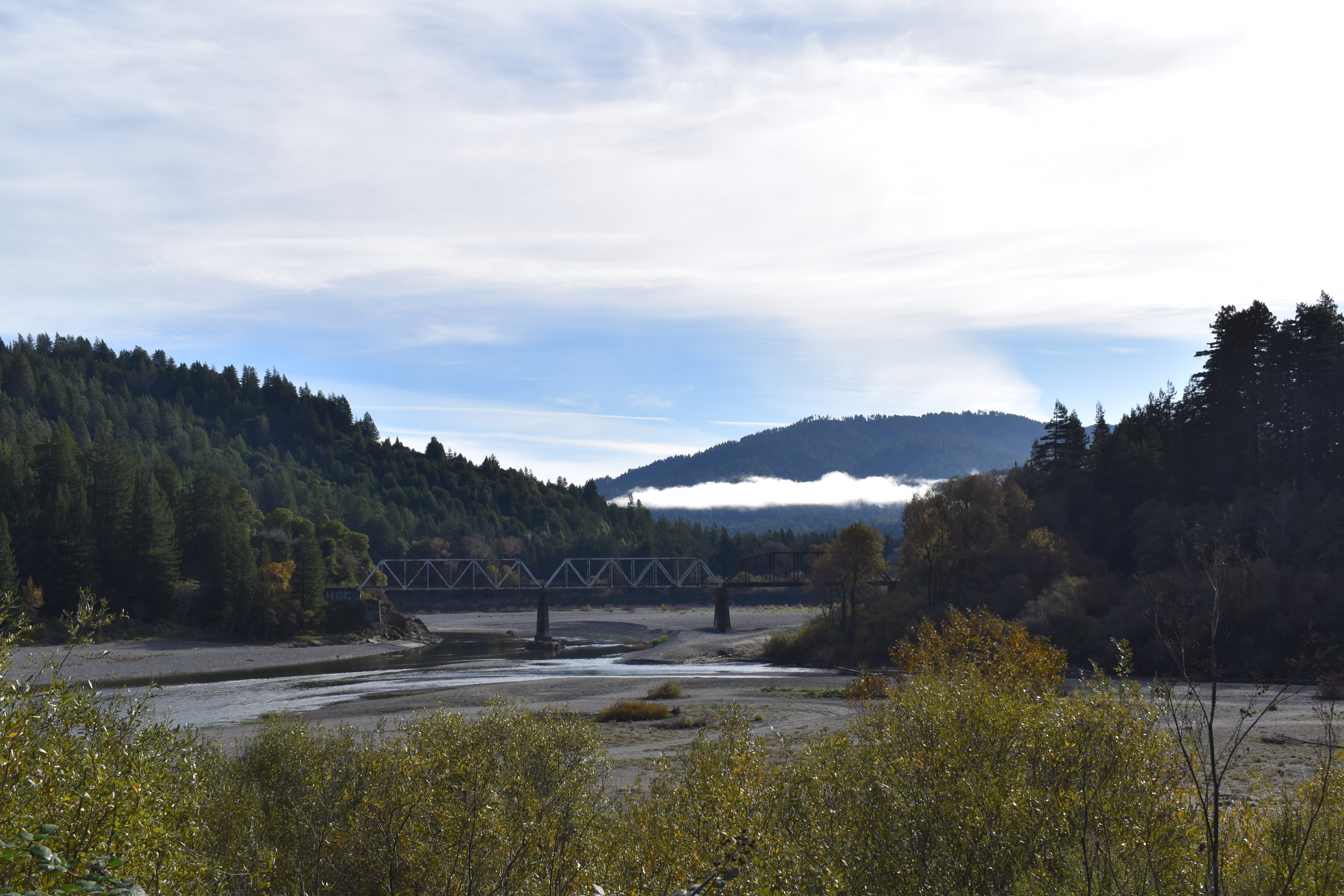 A view of a railroad bridge crossing the Eel River. There is mist on the mountains in the distance.